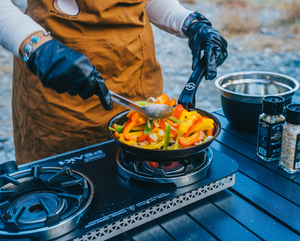 Person cooking peppers on a portable stove outdoors with a carbon steel pan